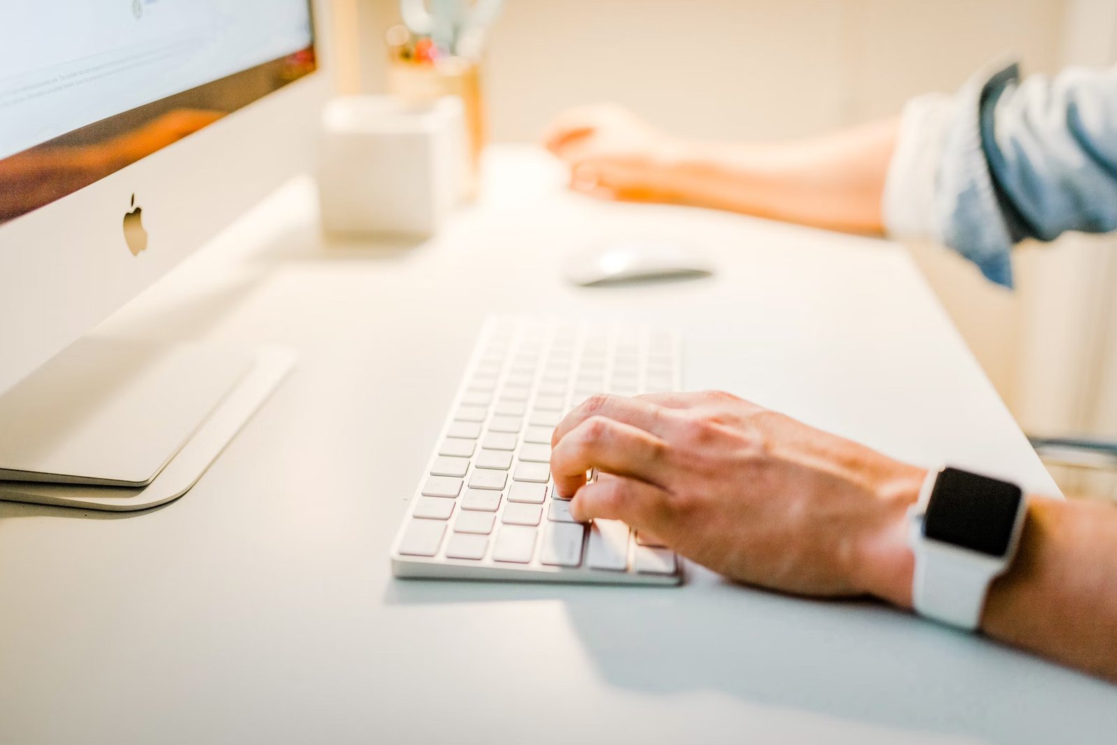 Hand typing on a keyboard next to an iMac and Apple Watch